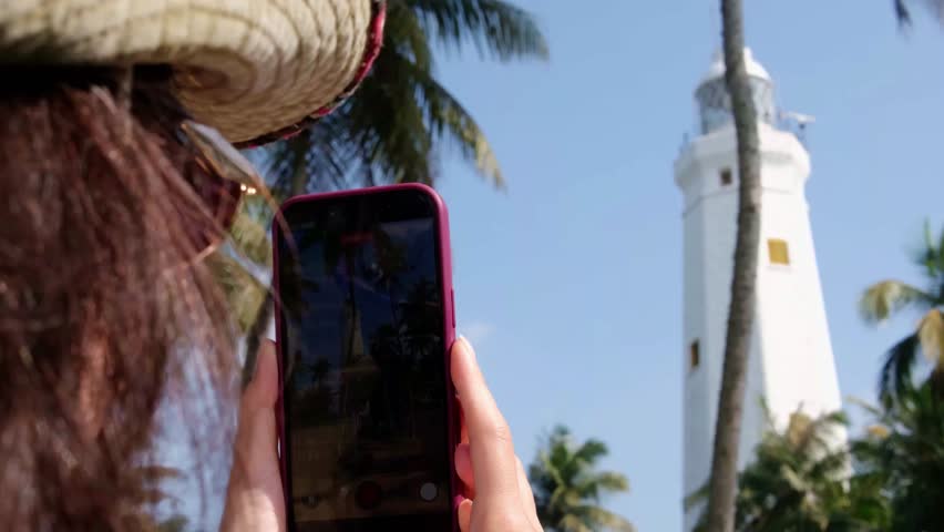 View of the white, tall lighthouse on the southern tip of the island of Sri Lanka. The lighthouse is surrounded by lush coconut trees.