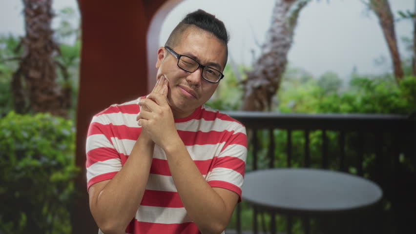 Man in red striped shirt wearing glasses with hand on cheek for toothache, wincing on a balcony outside a building surrounded by plants and a table; discomfort.