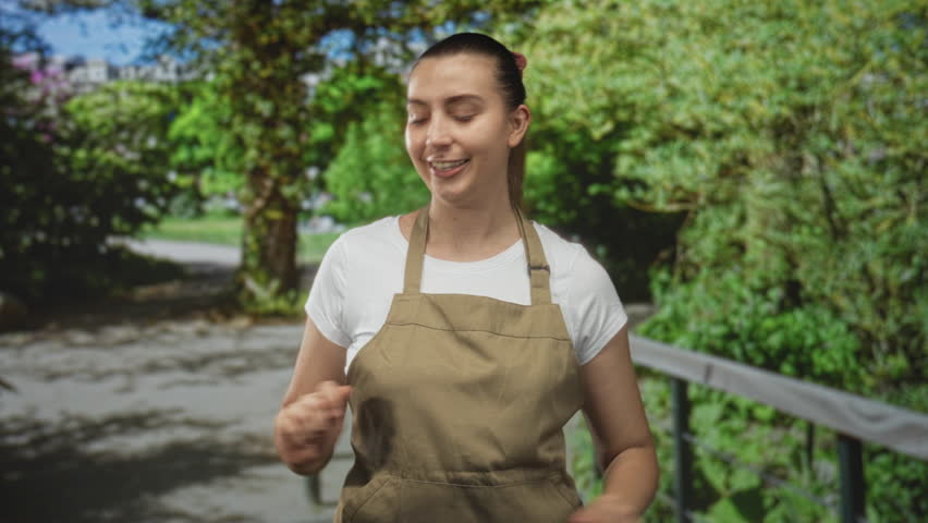 Woman in apron smiles while holding both palms up in a presenting gesture on a street lined with green foliage and railing; friendly welcome.