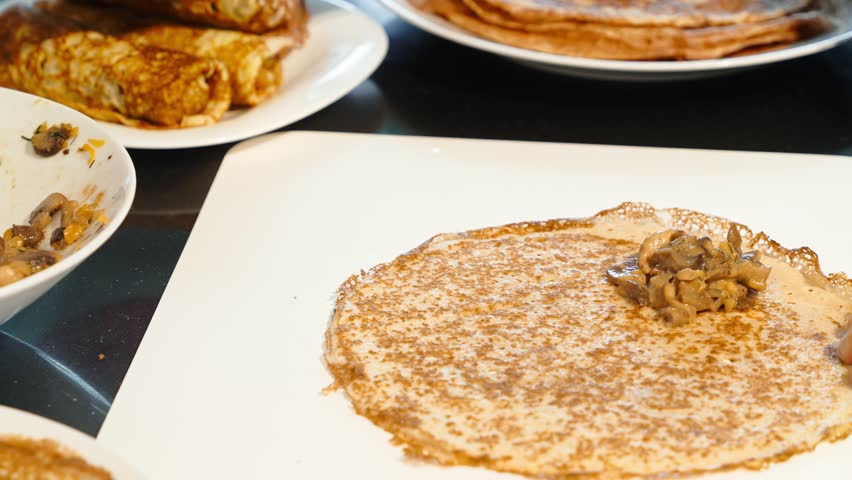 Person adds hot mushroom filling onto thin homemade crepe on white board and prepares to fold it. Stack of pancakes visible behind.