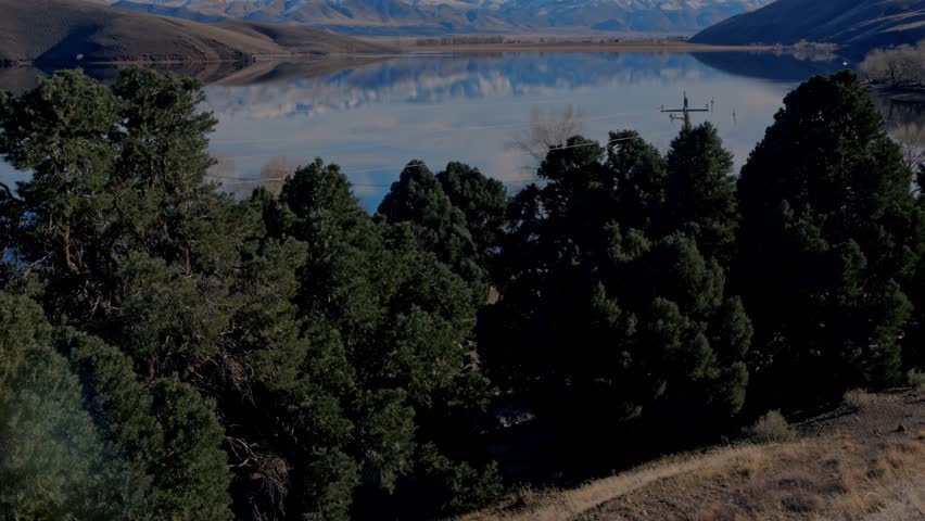Dense green pine trees frame perfect reflection of snowy Sierra Nevada mountains and rolling hills in calm blue Topaz Lake, cloudy sky, serene landscape at Nevada–California border.