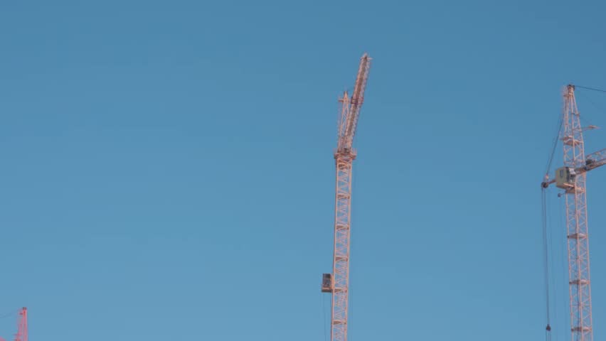 Construction cranes stand against a clear blue sky above a busy city