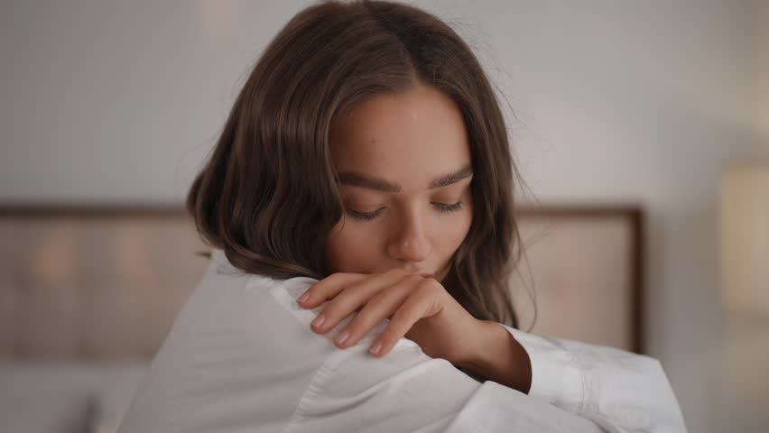Pensive young woman with natural makeup sitting on a bed and looking at the camera. Beautiful brunette female person posing in the bedroom