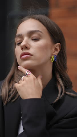 Beautiful young woman with natural makeup and stylish accessories posing outdoors. She thoughtfully touches her chin, then looks at the camera