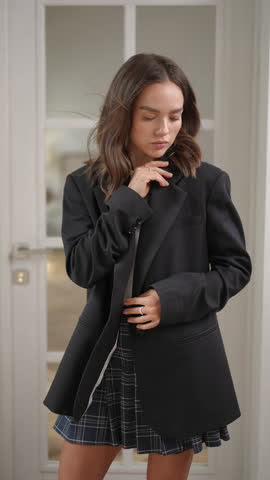 Beautiful young woman with brown hair posing indoors. She is wearing a trendy black oversized blazer and a plaid pleated mini skirt