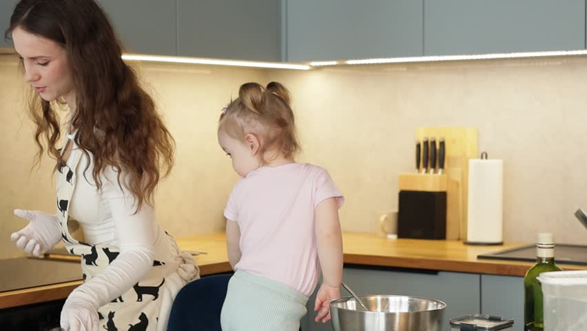 Young mother in cat print apron bakes with her toddler daughter in spacious modern kitchen with blue cabinets. Wide angle view of family cooking activity showing full kitchen interior.
