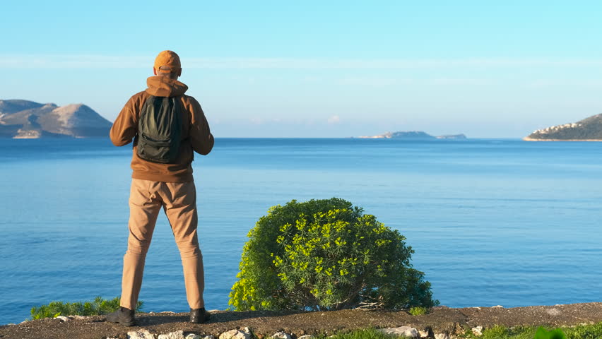 Traveler enjoying scenic ocean view with open arms. Lone backpacker standing on rocky coastal cliff, spreading arms wide while embracing panoramic ocean view with distant islands under bright sky