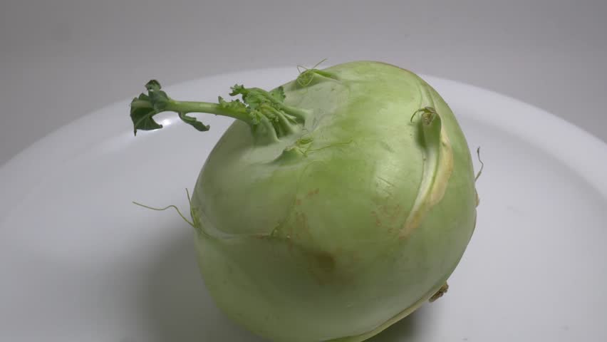 A fresh kohlrabi bulb with a small green leaf rotates slowly on a white plate in a studio setting, showcasing its unique shape and natural skin texture in close-up.