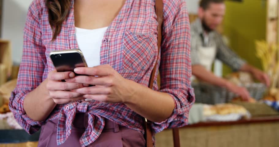 Woman typing phone at bakery, seeking likes while floating badges rising around face, baker working. Shopper, retail, bread, pastries, counter, rustic, cozy