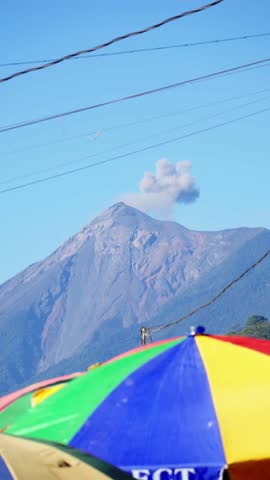 A Volcano Emits a Small Plume of Smoke Above a Colorful Market Umbrella - Vertical Shot