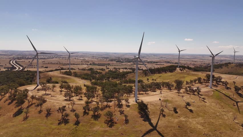 Aerial pullback across wind turbines rising from dry farmland with long shadows in afternoon light, Australia, Cullerin