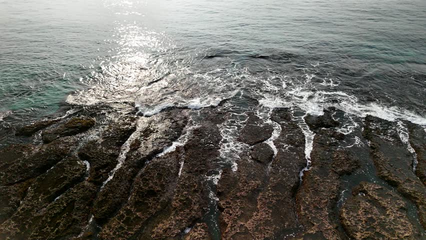 An aerial view of ocean waves splashing onto a rocky shoreline, showcasing the interplay between the sea and the land. The rugged rocks meet the dynamic water, creating a visually interesting landscape.