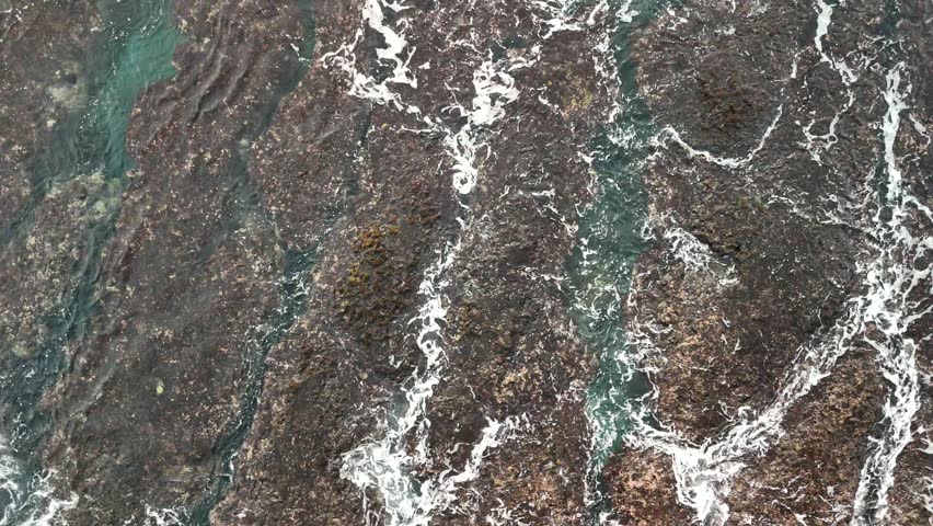 Aerial view of waves washing over a rocky shoreline. The image features dark, textured rocks and patches of algae (likely Phaeophyceae). The clear, turquoise water creates contrasting lines against the dark rocks, adding visual interest. Filmed with a drone.