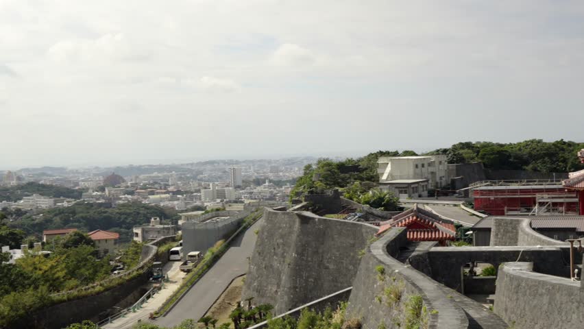 Panning shot captured from the historic Shurijo Castle walls overlooking the sprawling cityscape of Naha, Okinawa, Japan, under a diffused, cloudy winter sky.
