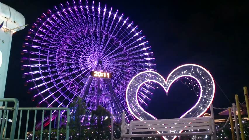 A mesmerizing nighttime photo of a giant urban Ferris wheel, likely the Cosmo Clock 21 in Yokohama, Japan, illuminated with vibrant purple LED lights, next to a large, white heart-shaped light installation. The image captures a romantic, festive atmosphere in a city amusement park setting