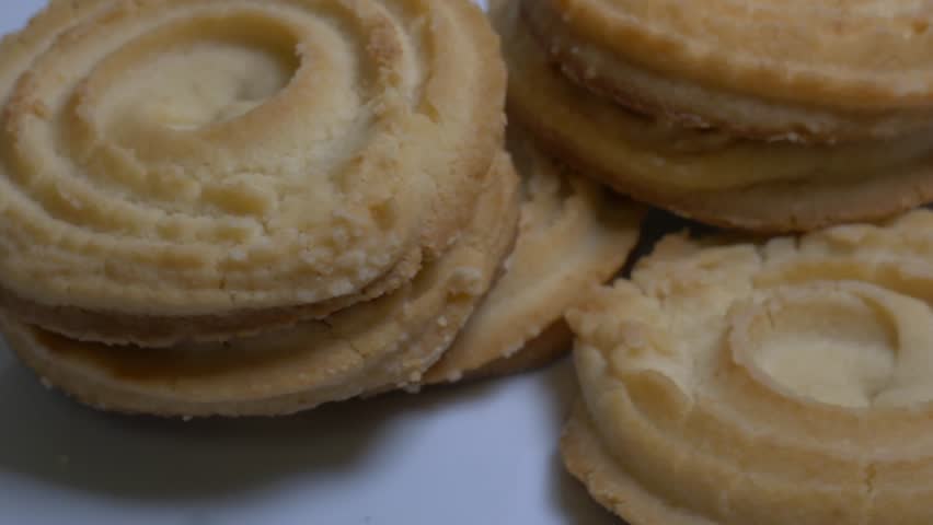A small stack of round butter cookies with a spiral pattern rotates slowly on a white plate, showcasing their crumbly texture and golden-baked edges in a studio setting.