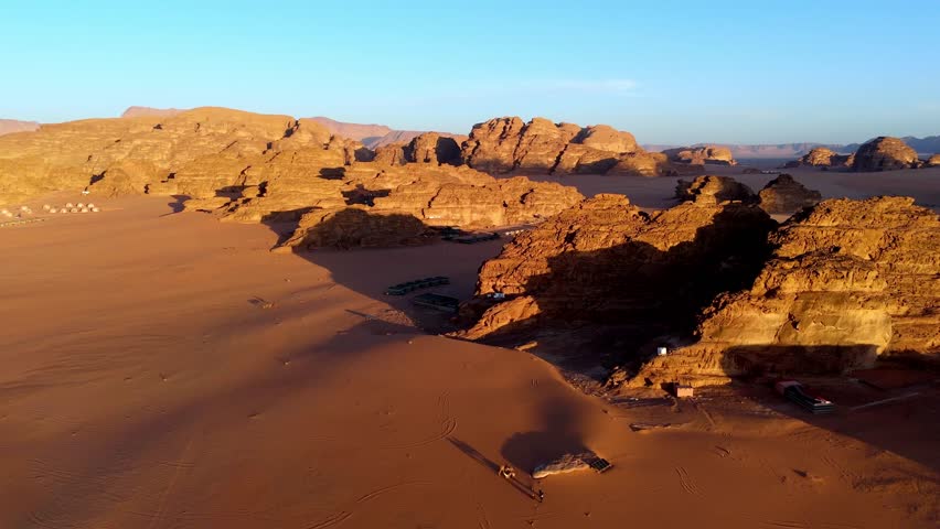 aerial shot capturing the warm glow of the sun setting over the rolling orange sand dunes and sandstone mountains of the Wadi Rum wilderness.