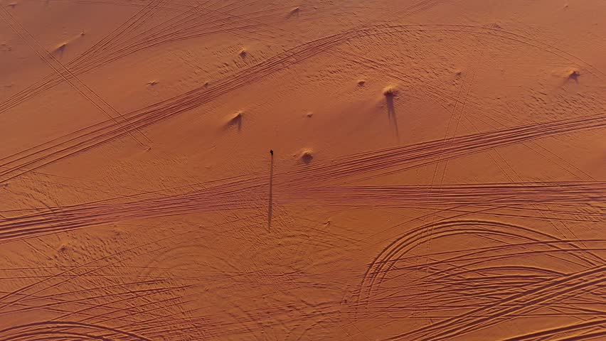 A stunning bird's-eye perspective of the Wadi Rum desert at sunset, where a lone individual walks across the vast orange sand, casting a dramatic long shadow amidst numerous tire tracks