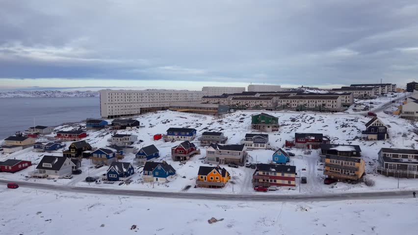 Cinematic aerial view of Nuuk, Greenland. Features a snow-covered houses, traditional houses, and an icy winter sea reflecting the sun under a dramatic sky.