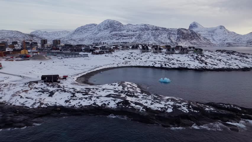 Drone shot of Nuuk, Greenland, featuring colorful houses on the snowy coast, a bright blue iceberg in the calm bay, and majestic Arctic mountains in the background during the winter season.