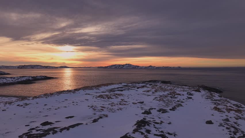 A breathtaking panoramic view of a golden sunset over a frozen Arctic coastline. Features snow-covered rocks, a calm icy sea reflecting the sun’s glow, and dramatic clouds in a winter sky.