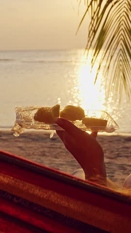 Vertical video. Woman Relaxing In Hammock Slowly Eating Fresh Pineapple While Watching Ocean Sunset Under Palm Trees Creating Peaceful Slow Life Vacation Mood.