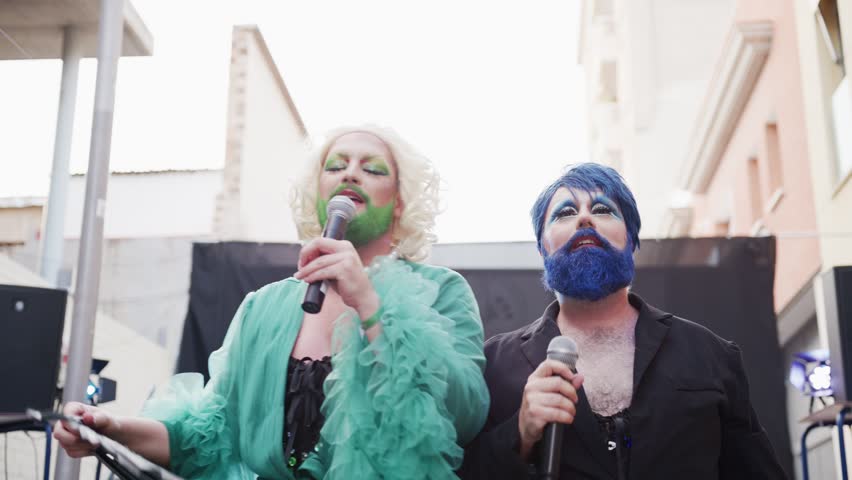 Two captivating bearded drag queens interact with the audience on stage in a lively courtyard. The colorful attire and enthusiastic crowd create an electric atmosphere at this unforgettable event.