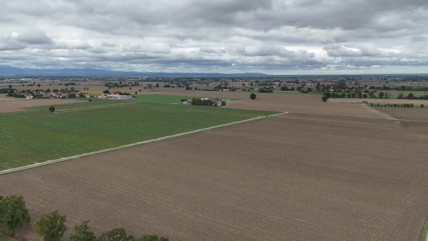 Expansive agricultural landscape with cultivated fields and scattered farmhouses under a dramatic cloudy sky near Piacenza, Italy