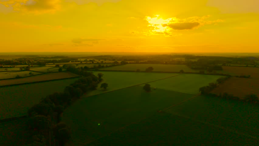 Radiant sunset lighting wide green farmland with golden glow. Warm evening sun descending over agricultural valley. Peaceful rural fields bathing in orange twilight