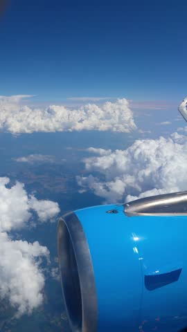 Aerial view from an airplane window over the Mediterranean, featuring a blue engine and wing flying above fluffy white clouds and land. Perfect for summer vacation and travel industry concepts