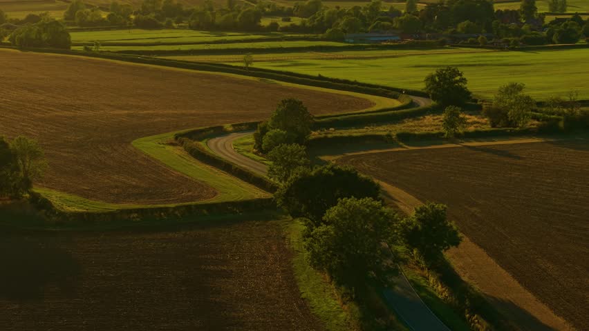Compact car driving along winding country road surrounded by sunset fields. Traveling car, highway driving, exploring countryside sunset. Small vehicle moving through rural curved roadway in golden