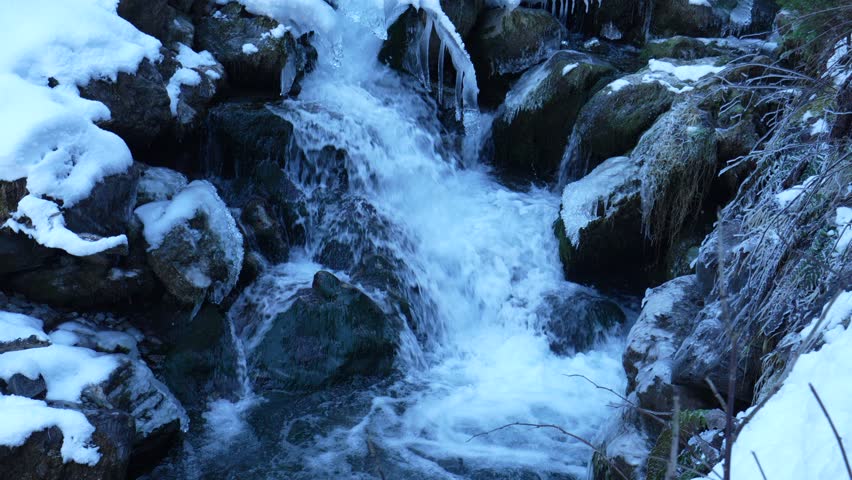 Cold mountain stream flowing through an icy and snowy winter landscape with frozen rocks