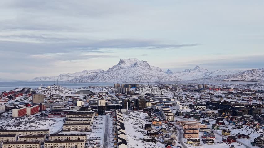 Scenic aerial panorama of Nuuk, Greenland, featuring colorful Nordic houses in a snow-covered landscape, modern city architecture, and the Arctic ocean under a soft winter sunset sky.