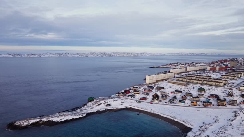 Scenic aerial panorama of Nuuk, Greenland, featuring colorful Nordic houses in a snow-covered landscape, modern city architecture, and the Arctic ocean under a sunset sky.