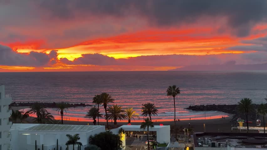 Vibrant orange sunset over the Atlantic with palm trees, calm waves, and a coastal resort beach, viewed from above. Dramatic clouds and reflections create a tropical travel mood with copy space