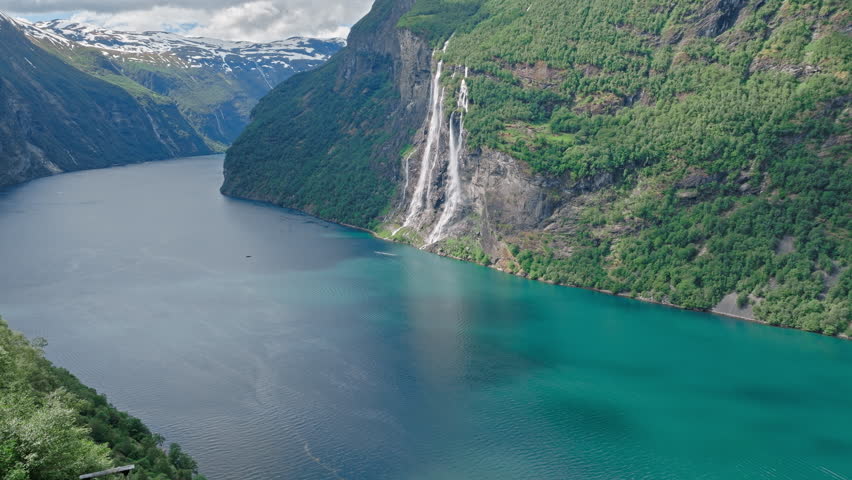 Wide aerial view of Geirangerfjord winding between steep mountains in Norway. Waterfalls and forests define this classic Norwegian fjord scene.