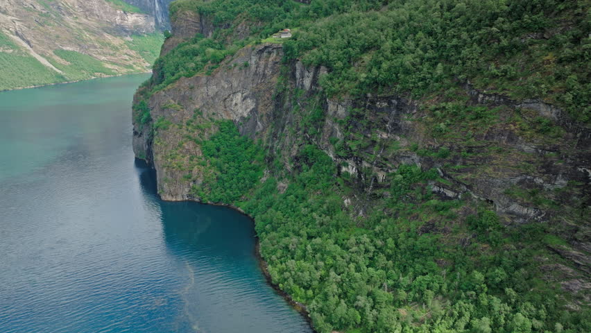 Aerial movement past towering cliffs rising directly from the fjord in Norway. The scene highlights the raw scale of Norwegian landscapes and deep blue water.