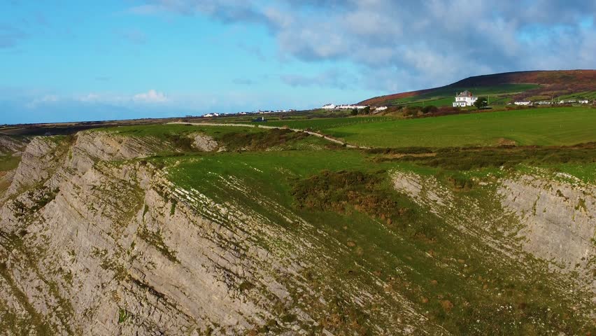 Descending Aerial with Lone House on Hilltop with Steep Cliffs with Stone Wall Lining Coast Path with Small Village in Background. Travel Nature Drone Clip. Beautiful Welsh Gower Peninsula Coast.