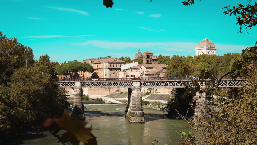 Wide view of a historic river bridge and classic cityscape in Rome, Italy. Urban landscape with European architecture, green trees and calm river under a clear blue sky. No people.