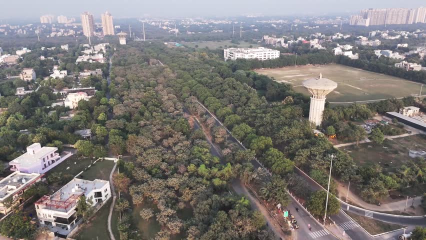 Aerial view of Greater Noida showing a water tower rising above dense green cover, with residential high-rises in the distance and light haze revealing the city’s expanding urban landscape.