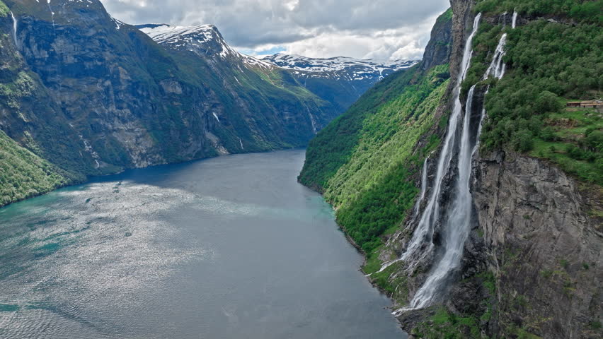 Extended aerial view following the fjord as it curves between towering cliffs and waterfalls in western Norway. The Norwegian scenery appears vast and serene under soft daylight.