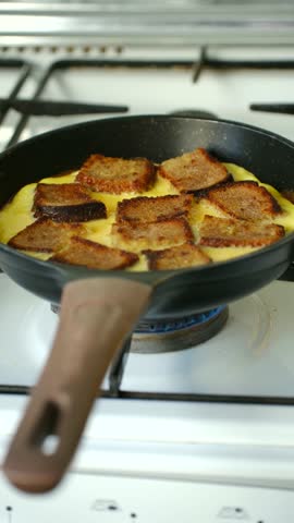 Delicious and hearty french toast omelette cooking in a black frying pan over a vibrant blue flame from a gas stove burner, showcasing a simple yet satisfying homemade breakfast recipe. Vertical