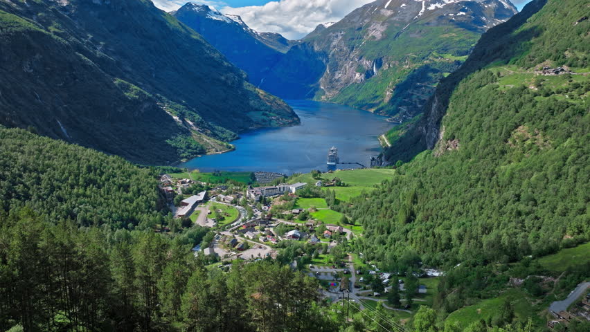 Aerial view of a Norwegian fjord village framed by steep mountains and deep blue water. Clear daylight reveals compact buildings nestled between forest and shoreline.
