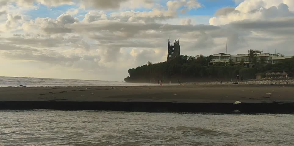 This image appears to be a screenshot of a “Video details” page on a mobile device. In the center, there is a wide cinematic scene of a beach during what looks like late afternoon or early evening. The sky is filled with soft, golden clouds, suggesting sunset light.
In the distance, on a small hill overlooking the shoreline, there is a tall, dark, castle-like building with two pointed towers. Surrounding it are several lighter-colored buildings partially hidden by trees. The beach in the foreground looks broad and calm, with gentle waves near the shore and a few small figures of people walking along the sand. The overall atmosphere feels peaceful and slightly dramatic due to the lighting and the silhouette of the tall structure against the sky.