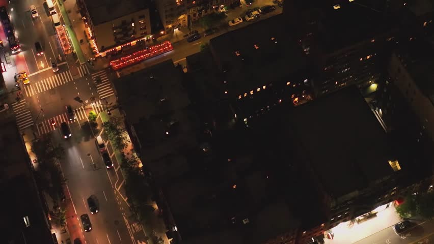 A busy street in New York at night. Cars move along the roads while streetlights and building lights create a vibrant city atmosphere. The view is from above.