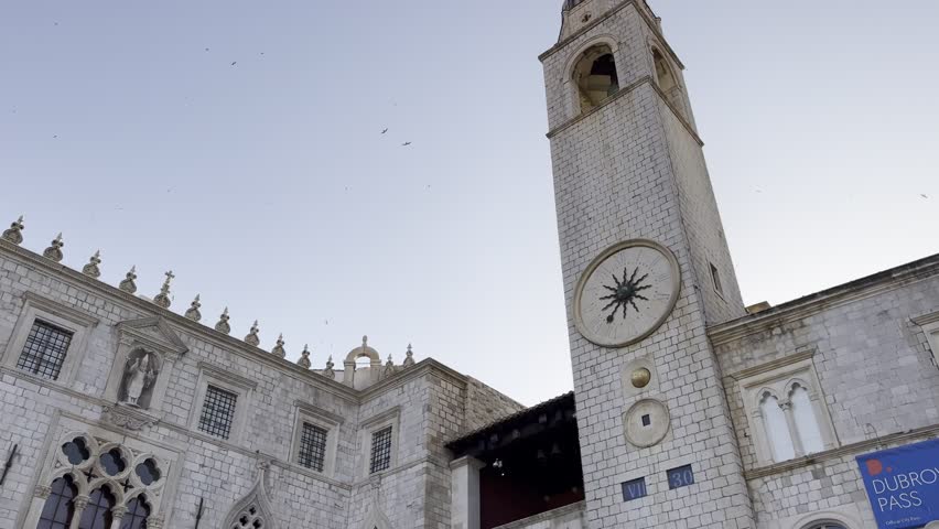 Slow cinematic rotating shot of the Clock tower of Dubrovnik during early morning light with starlings flying across a clear sky and historic stone architecture in the old town