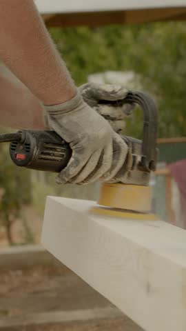 Vertical Screen: A worker uses an orbital sander on wooden beams to prepare them for finishing. The outdoor setting includes greenery and construction materials in the background.