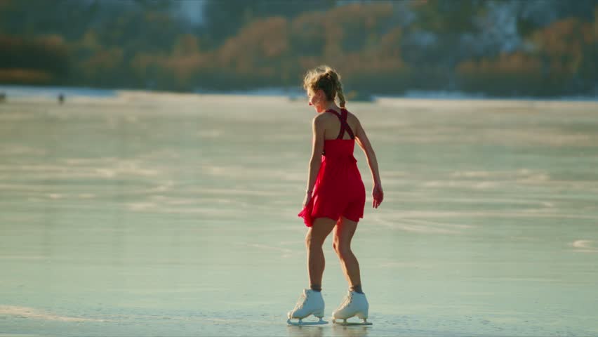 A young girl gracefully skates across the glistening ice of a frozen lake, adorned in a vibrant red dress and ice skates, showcasing her agility and joy in the serene winter landscape.