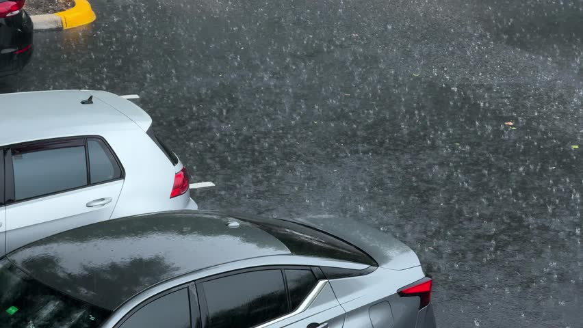 Heavy rain pours down on cars parked in a wet asphalt parking lot. Raindrops splash on the dark pavement, creating a dramatic weather scene and low visibility for drivers.