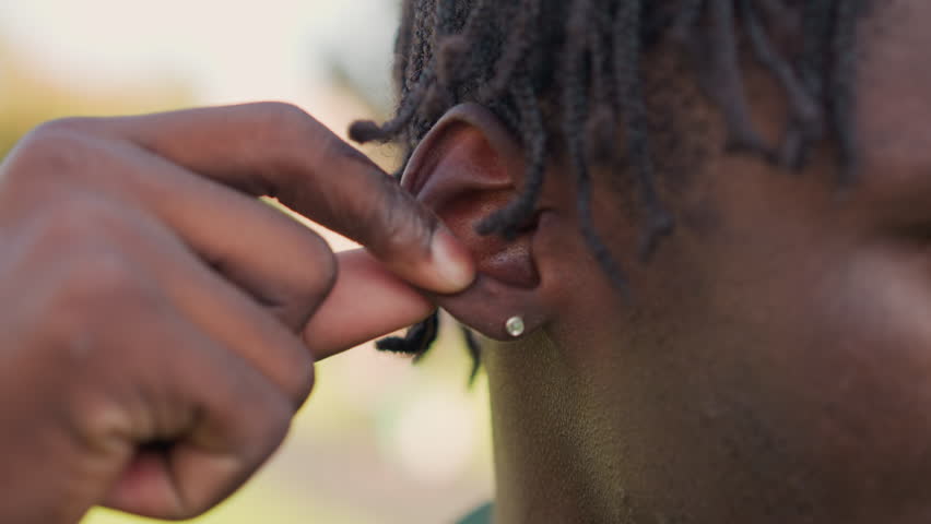 Black Soldier Placing Earbud In Ear With Careful Fingers, CloseUp Of Earring, Locs And Attentive Eyes Outdoors Scene Shows Connection, Private Communication And Readiness To Listen Or Speak